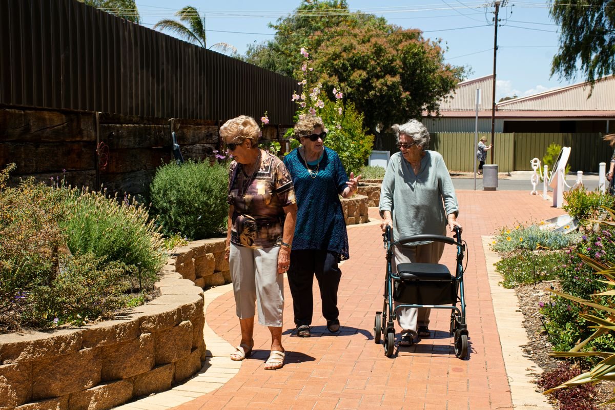 Three older women walking and talking together along a sunny garden path